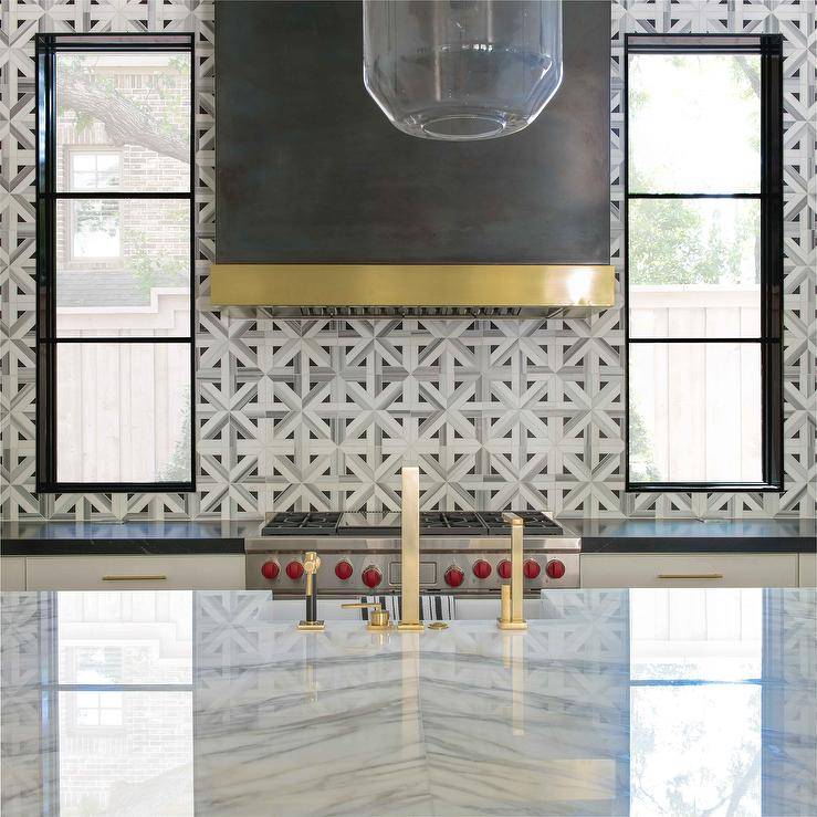 Black dome range hood with brass trim brings a striking accent to a contemporary kitchen cooktop finished with a white and gray marble geometric backsplash.