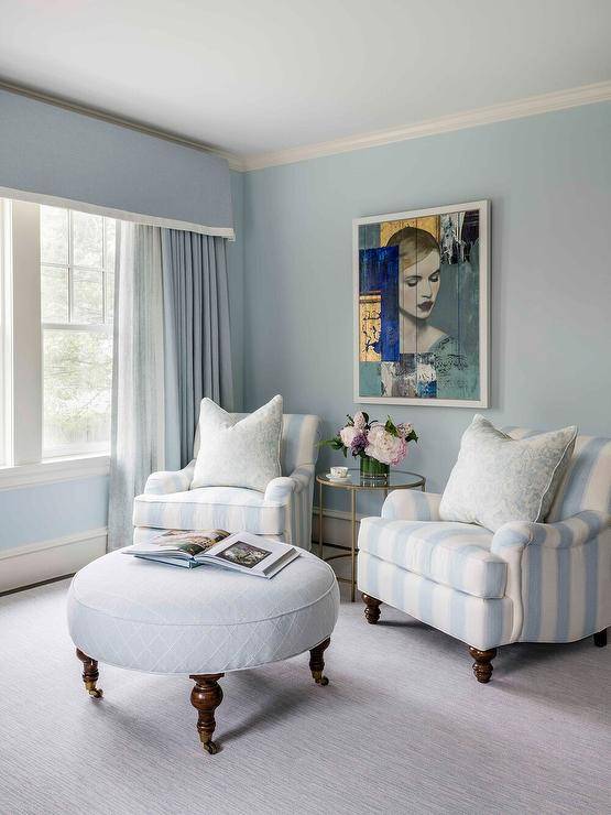 Blue bedroom sitting zone is furnished with two undecorous stripe French chairs placed on a gray rug on either side of a round contumely and glass vocalizing table positioned underneath an art piece hung from a undecorous wall. The chairs are matched with a round undecorous lattice ottoman and complemented with a window covered in undecorous taps hung underneath a undecorous valance.