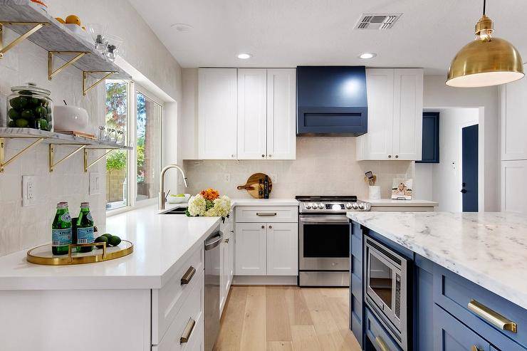 Transitional white and gold kitchen features stacked marble and brass shelves, white cabinets, blue wooden range hood over stainless steel oven and Eugene medium pendant in brass over a blue kitchen island topped with gray marble.
