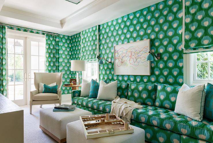 Bold blue and green print wallpaper complement a green and blue sofa accented with ivory and green pillows and matched with two ivory ottomans and a cream wingback chair. An abstract art piece hangs above the sofa between green sconces.