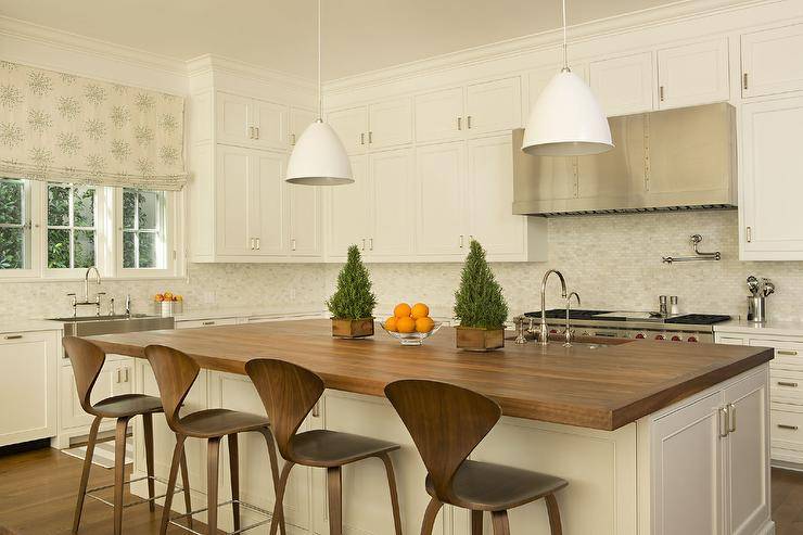 Two white industrial pendants are hung over an ivory kitchen island accented with a butcher block countertop. Cherner Stools are placed at the island facing a stainless steel sink with a polished nickel gooseneck faucet.