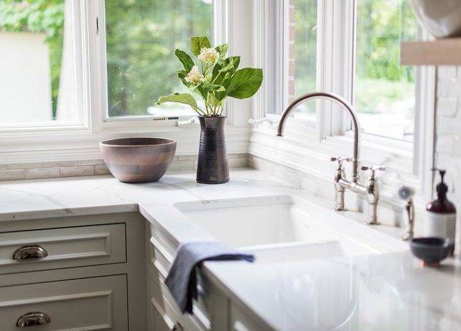 Beneath windows, a cast iron dual sink matched with a polished nickel deck mount faucet is fitted over white kitchen cabinets donning polished nickel cup pulls and a white quartz countertop.