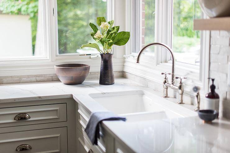 Beneath windows, a cast iron dual sink matched with a polished nickel deck mount faucet is fitted over white kitchen cabinets donning polished nickel cup pulls and a white quartz countertop.