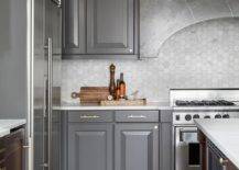 Kitchen features charcoal gray cabinets with a dark gray marble curved range hood over honed light gray hexagon marble backsplash tiles and reeded walls.