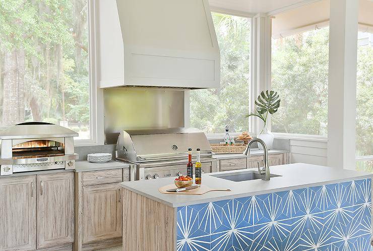 Outdoor kitchen features an island with blue concrete tiles, a concrete countertop with sink, a range hood over grill and a pizza oven.