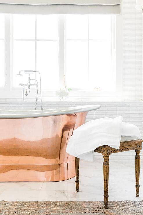 A polished nickel tub filler is mounted to a ledge beneath a window and paired with a French copper bathtub positioned on marble floor tiles.