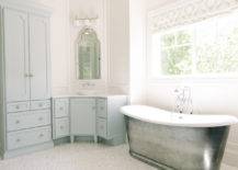 Blue bathroom features a cast iron tub and a vintage style tub filler placed under windows dressed in a white and grey geometric roman shade. Master bathroom boasts a blue corner washstand painted Benjamin Moore Woodlawn Blue topped with white marble under an Allen + Roth Hovan Arch Frameless Mirror illuminated by a three light sconce alongside a mosaic marble floor.