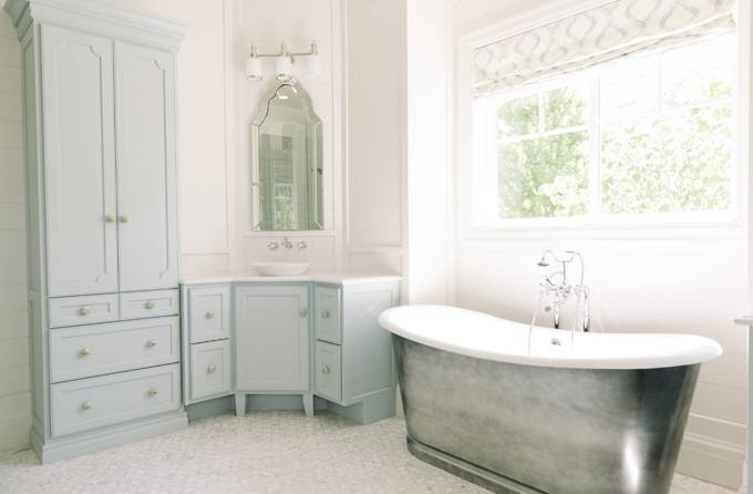 Blue bathroom features a cast iron tub and a vintage style tub filler placed under windows dressed in a white and grey geometric roman shade. Master bathroom boasts a blue corner washstand painted Benjamin Moore Woodlawn Blue topped with white marble under an Allen + Roth Hovan Arch Frameless Mirror illuminated by a three light sconce alongside a mosaic marble floor.