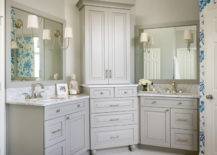Beautiful bathroom features gray his and hers vanities topped with white marble under gray mirrors illuminated by Reed Single Sconces flanking corner center console cabinet stacked over gray dresser.