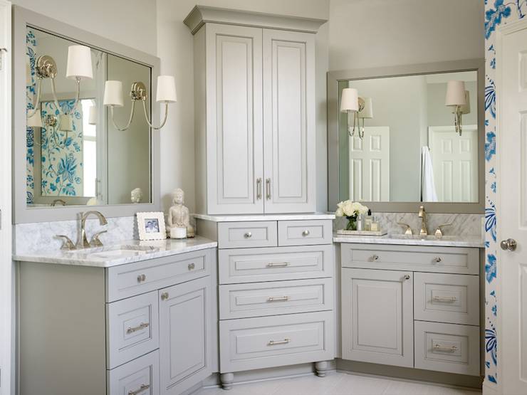Beautiful bathroom features gray his and hers vanities topped with white marble under gray mirrors illuminated by Reed Single Sconces flanking corner center console cabinet stacked over gray dresser.