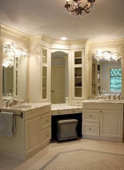 Beautiful master bathroom design with gray vanity ottoman tucked under corner vanity accented with inset mirror and glass-front cabinets flanked by his and her vanities featuring ivory cabinets paired with white marble countertops and sconces mounted on inset mirrors.