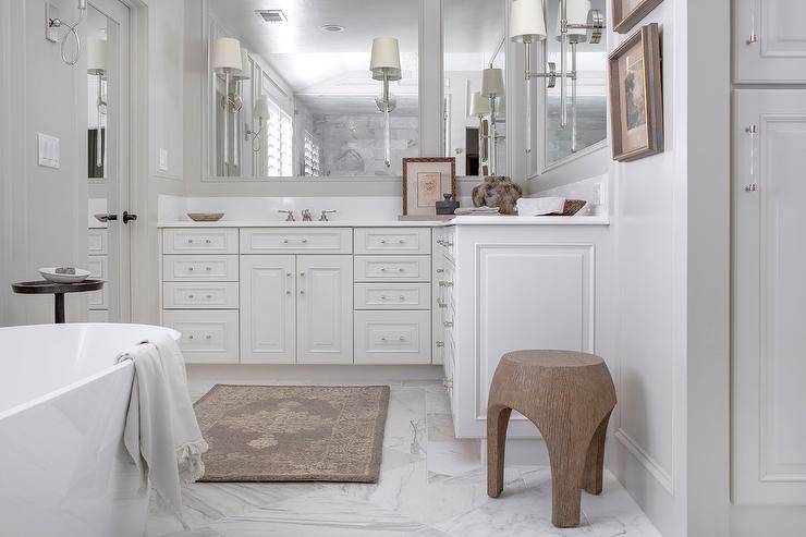 An ivory and gray rug sits on marble floor tiles in front of a white l-shaped washstand adorned with nickel and glass pulls and lit by Camille Long Sconces mounted to white framed vanity mirrors.