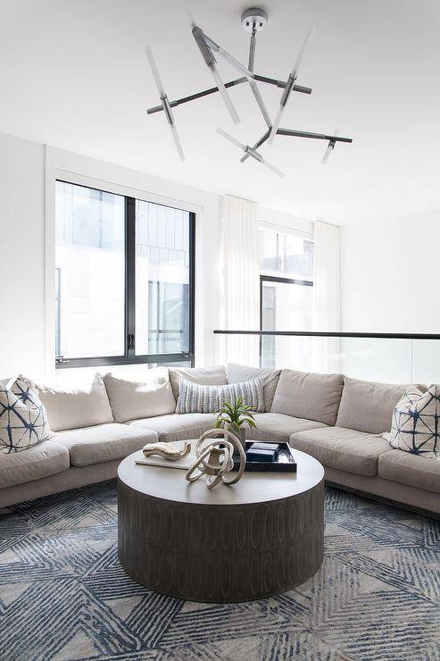 In front of a glass pony wall, a tan corner sectional accented with tan and blue pillows sitso n a tan and blue wool rug facing a round black coffee table.