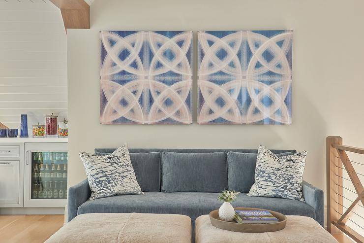 Two pink and blue canvas art pieces hang over a blue velvet sofa topped with white and blue pillows and positioned facing side-by-side cream wool ottomans.