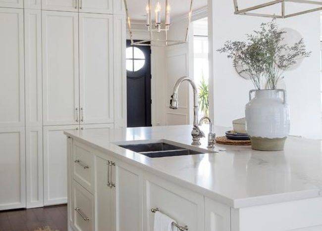Two Darlana Lanterns hang over a white center island fitted with a stainless steel dual sink and a polished nickel gooseneck faucet. The island is finished with a concealed dishwasher hidden behind a white paneled door.