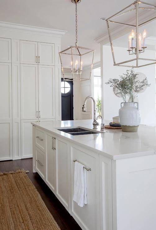 Two Darlana Lanterns hang over a white center island fitted with a stainless steel dual sink and a polished nickel gooseneck faucet. The island is finished with a concealed dishwasher hidden behind a white paneled door.