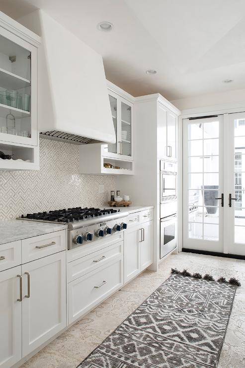 Kitchen features white cabinets with white and tan diamond pattern backsplash tiles and a white range hood.