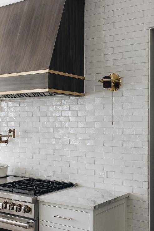 Kitchen features a dark brown wooden range hood with brass straps on white wall tiles lit by a brass sconce.