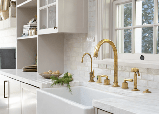 Kitchen features light gray cabinetry accented with brass pulls, a brass gooseneck faucet with a farmhouse sink and glazed white subway tiles.