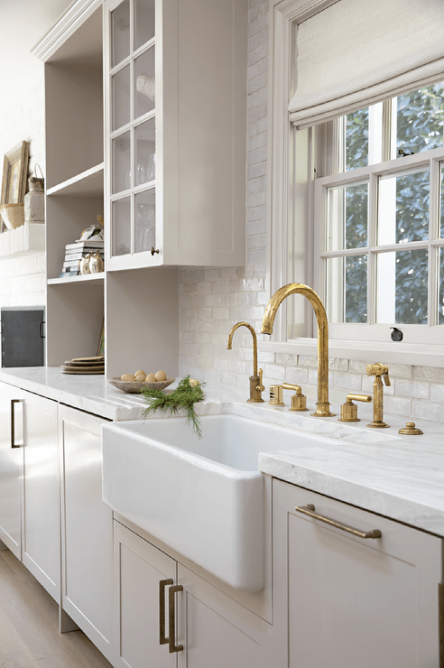 Kitchen features light gray cabinetry accented with brass pulls, a brass gooseneck faucet with a farmhouse sink and glazed white subway tiles.