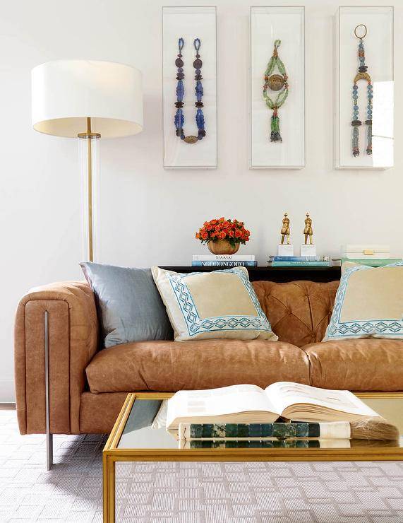 Contemporary living room design features a modern brown leather tufted sofa accented with cream, blue and silver pillows, a gold leaf coffee table over a tan rug, a colorful art gallery over the sofa and a brass and cream floor lamp.