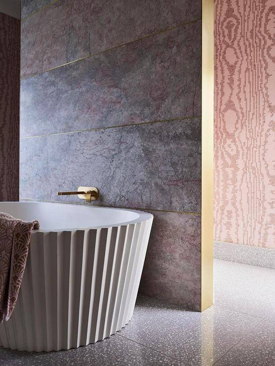 A brass tub filler is fixed to a pink and gray marble wall over a fluted freestanding bathtub placed on gray recycled floor tiles.