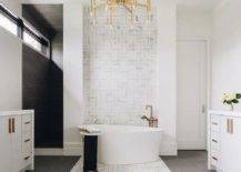 A brass chandelier illuminates a luxurious bathroom featuring an oval freestanding bathtub with a catty corner aged brass floor mount tub filler placed on marble and brass basketweave accent floor tiles and in front of marble and brass basketweave wall tiles.
