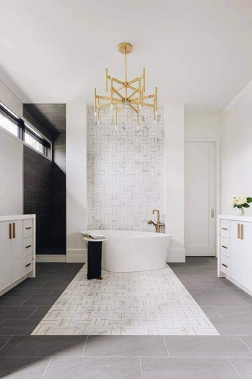 A brass chandelier illuminates a luxurious bathroom featuring an oval freestanding bathtub with a catty corner aged brass floor mount tub filler placed on marble and brass basketweave accent floor tiles and in front of marble and brass basketweave wall tiles.