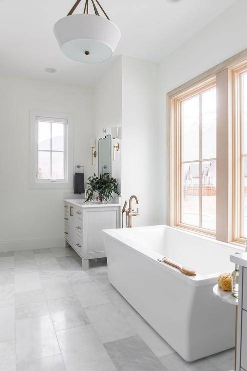 A rectangular freestanding bathtub with a polished nickel floor mount tub filler sits beneath a window on and between gray his and hers washstands.