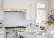 A light blue and gold French stove is flanked by inset white shaker cabinets donning with brass pulls and fixed beneath a white range hood lined with brass trim and positioned between by stacked cabinets.