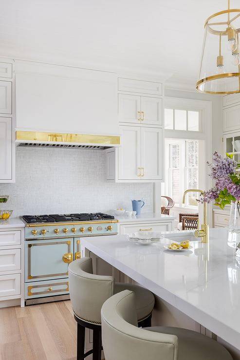 A light blue and gold French stove is flanked by inset white shaker cabinets donning with brass pulls and fixed beneath a white range hood lined with brass trim and positioned between by stacked cabinets.