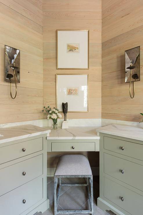 Two gold framed art pieces hang from a tan plank wall over a green corner makeup vanity topped with a honed white marble countertop paired with a gray counter stool placed on gray floor tiles. The vanity is flanked by green washstands accented with satin nickel knobs and lit by mirrored sconces.