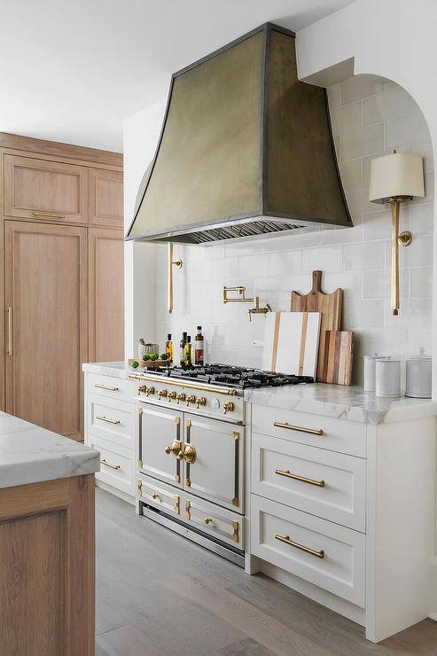 A cooking alcove is filled with a white La Cornue CornuFé 110 Range paired with glossy white tiles and brass sconces alongside a verdigris hood.