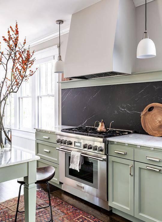 A gray range hood is fixed over a soapstone slab cooktop backsplash mounted over a stainless steel oven range flanked by green cabinets finished with nickel hardware and a white marble countertop.