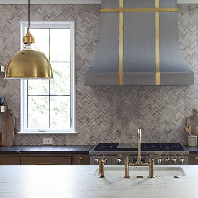 A stainless steel French range hood with brass straps is fixed against gray marble cooktop tiles between windows and over a stainless steel oven range. The oven range sits between brown oak cabinets donning brass hardware a black marble countertop.