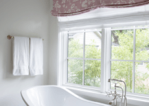 A white freestanding high-curve back tub pairs with a sleek polished nickel faucet offering a quiet place to soak, beside a window with a pink floral roman shade. A small brass sputnik chandelier brings a finishing touch of mod over the tub and white mosaic floor tiles.