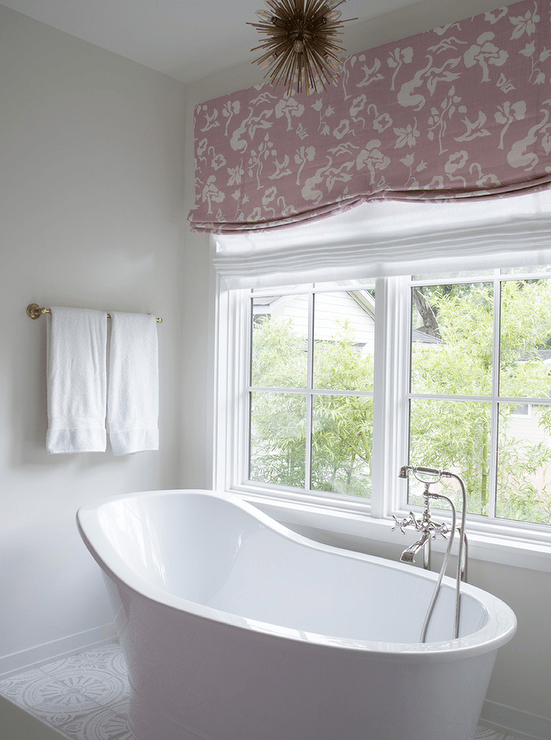A white freestanding high-curve back tub pairs with a sleek polished nickel faucet offering a quiet place to soak, beside a window with a pink floral roman shade. A small brass sputnik chandelier brings a finishing touch of mod over the tub and white mosaic floor tiles.