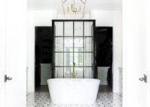 Luxurious bathroom boasts white, gray, and black hexagon floor tiles leading to an oval freestanding bathtub matched with a nickel floor mount tub filler and lit by a brass lantern. The tub is located in front of a glass and steel walk-in shower enclosure.