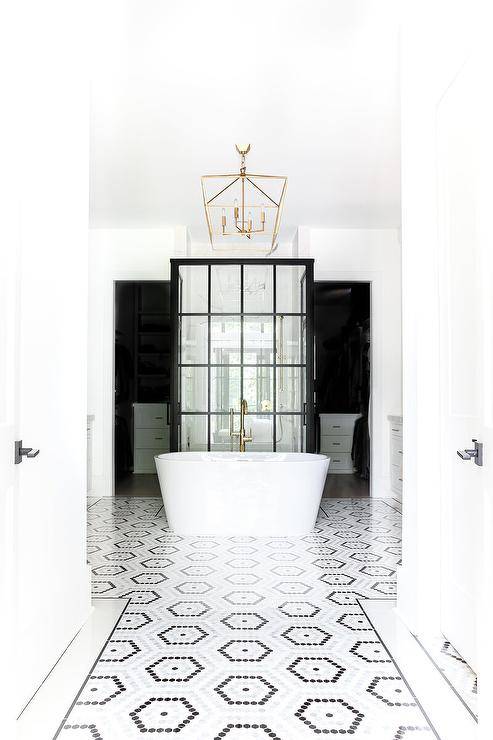 Luxurious bathroom boasts white, gray, and black hexagon floor tiles leading to an oval freestanding bathtub matched with a nickel floor mount tub filler and lit by a brass lantern. The tub is located in front of a glass and steel walk-in shower enclosure.