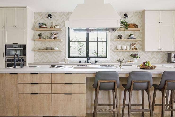 Spacious kitchen design features gray leather stools at a beige veneer center island with cooktop under a white range hood and floating shelves on herringbone tiles.