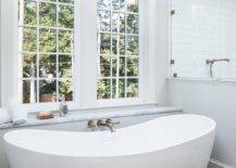 Brushed gold wall mount tub filler designed with an oval freestanding tub illuminated with a brass hanging light in a transitional bathroom design. A window over the tub adds lovely natural light to an already bright and airy bathroom design.