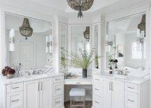 Hung from a vaulted ceiling over wood-like floor tiles, a gray beaded chandelier lights a well-appointed bathroom. A chrome vanity stool sits beneath a drop-down makeup vanity fitted in a corner and topped with a white quartz countertop mounted beneath white framed vanity mirrors. The makeup vanity is located between white shaker washstands boasting chrome pulls and white quartz countertops finished with chrome faucets mounted under white frame vanity mirrors.