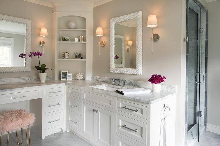 A pink faux fur stools sits at a white built-in makeup vanity accented with a gray and white marble countertop and polished nickel pulls. Above the vanity a white mirror is lit by Camille Long Sconces, while styled shelves are mounted in the corner. On an adjacent wall, a white washstand is finished with a polished nickel faucet kit fixed to a gray and white marble countertop beneath a white mirror flanked by Camille Long Sconces.