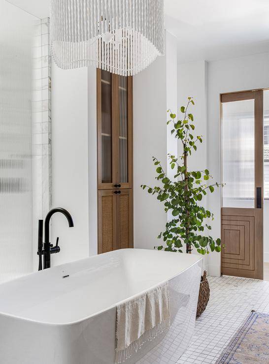 A white beaded chandelier illuminates a freestanding bathtub with an oil rubbed bronze floor mount tub filler and marble grid floor tiles.