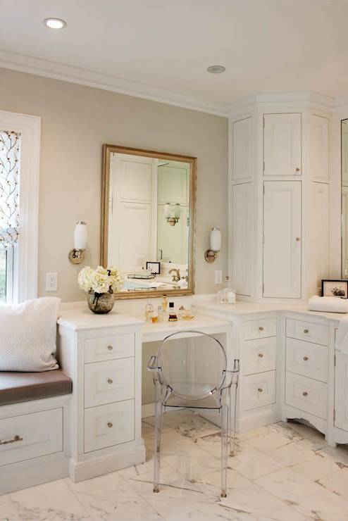 Stunning bathroom with built-in window seat to the left of a next to a drop-down vanity lined with a Ghost Chair. A large gilt mirror flanked by nickel and white glass sconces hangs above the vanity with a corner cabinet finishing the space over marble tiled floors.