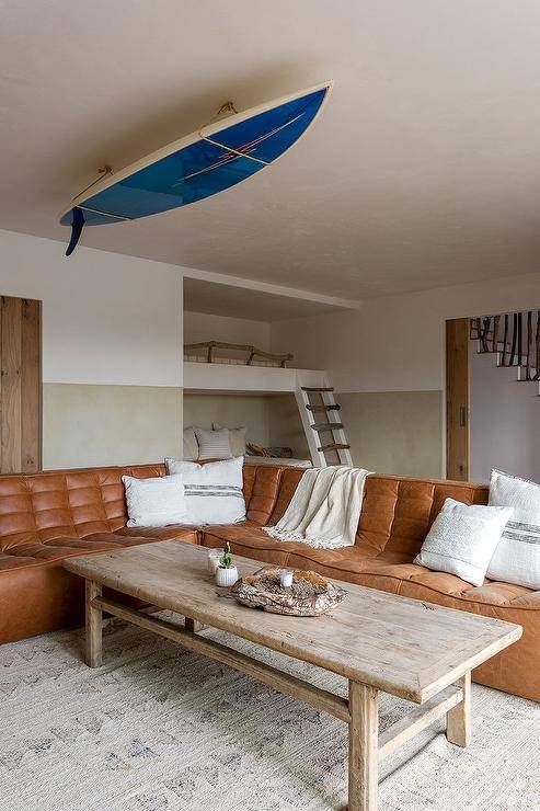 Living room features a brown leather tufted sectional with a reclaimed wood coffeet table under a decorative ceiling surfboard.