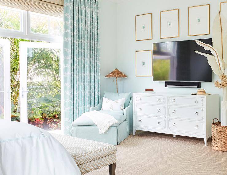 Art surrounds a television placed on a white Greek key dresser in a bedroom self-aggrandizing a undecorous chaise lounge positioned in a corner in front of glass doors covered in undecorous taps layered over bamboo roman shades.