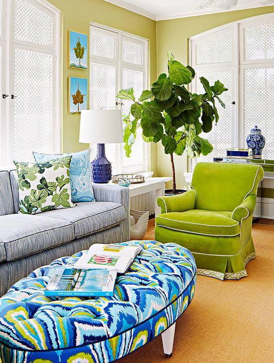Contemporary green and blue living room features an oval green and blue tufted ottoman placed in front of a blue striped sofa accented with blue and green pillows. Stacked art pieces hang from a green wall over a white Parsons side table illuminated by a cobalt blue lamp, as a fiddle leaf fig plant sits next to a green velvet roll arm chair.