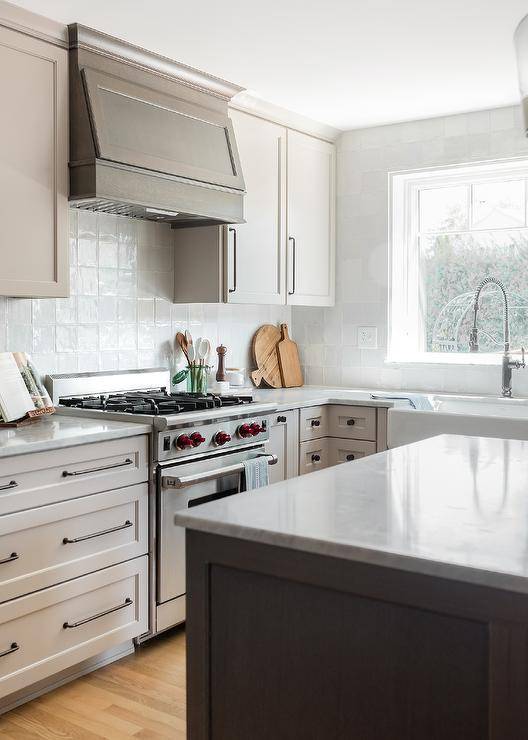 Kitchen features a brown wooden kitchen range hood and gray glazed grid tiles over a wolf range, flanked by with light taupe cabinets with oil-rubbed bronze pulls and a marble topped kitchen island.