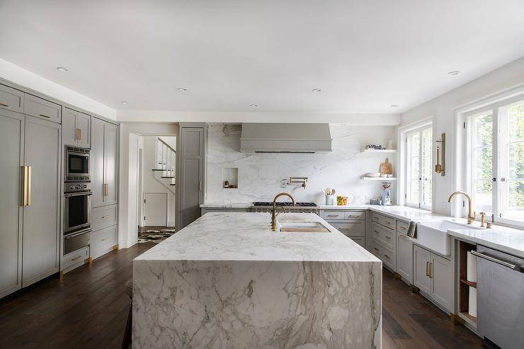 A sliding cutting board accents a kitchen sink with brass gooseneck faucets mounted to a marble waterfall edge island.